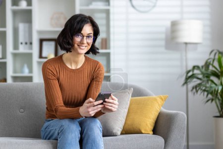 Casual woman sitting comfortably on a couch at home, deeply engaged in using her smartphone in a well-lit, cozy living room.