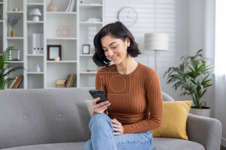 Eine fröhliche junge Frau, die mit ihrem Smartphone beschäftigt ist, sitzt gemütlich auf einem modernen Sofa in einem gut beleuchteten Wohnzimmer.