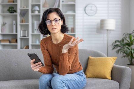 Young woman sitting on sofa, looking confused while reading a text message that seems to deliver bad news.