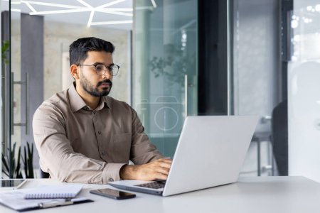 Konzentrierter indischer Mann in schicker Freizeitkleidung, der ernsthaft arbeitet und denkt, während er einen Laptop in einer modernen Büroeinrichtung benutzt.