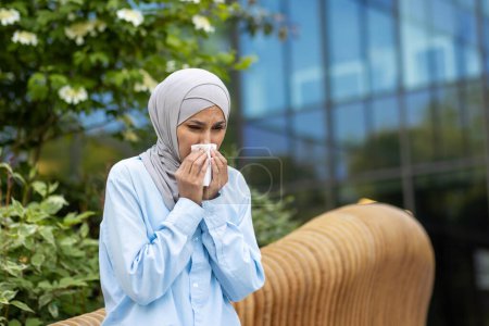 A young woman in a blue hijab feeling unwell, sneezing into a tissue in an urban park setting with greenery.