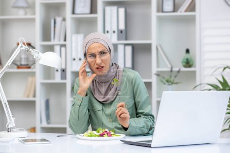 Exhausted muslim woman in hijab sitting at desk and eating salad while working on laptop. Tired female experiencing headache in office with white shelves and decorative items.