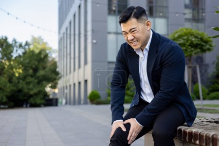Photo for Contorted in pain, a young Asian businessman sits on a bench near an office center and holds his knee with his hands. Got an injury, inflammation. - Royalty Free Image