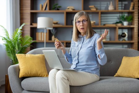 Reife Frau zu Hause schaut verwirrt auf Kreditkarte, sitzt mit ihrem Laptop auf der Couch in einem modernen Wohnzimmer-Ambiente.