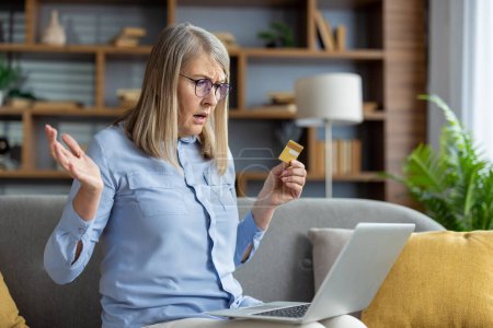 Ältere Frau sitzt verwirrt mit Kreditkarte und Laptop auf der Couch und beschäftigt sich möglicherweise mit Online-Shopping oder Bankgeschäften.
