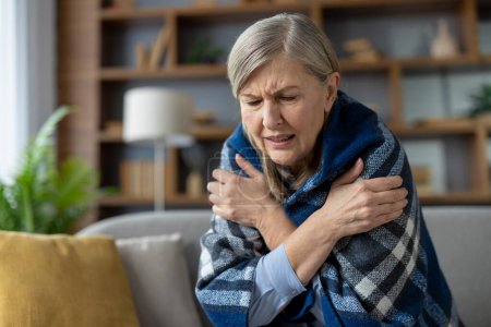 Une femme âgée souffrant de frissons, assise sur le canapé enveloppé dans une couverture confortable, visiblement froide et inconfortable.