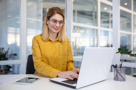 Selbstbewusste und ermächtigte Berufstätige in einem gelben Hemd lächeln, während sie in einem hellen Büro an einem Laptop arbeiten.