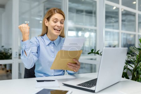 Téléchargez les photos : Une employée de bureau joyeuse célèbre le succès avec une enveloppe dans un espace de travail moderne, évoquant des sentiments de réussite et de bonheur. - en image libre de droit