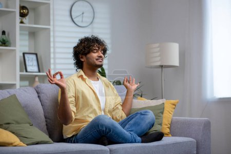 Photo for Peaceful Indian man with curly hair meditating in lotus pose on a sofa at home, exhibiting calmness and relaxation. - Royalty Free Image