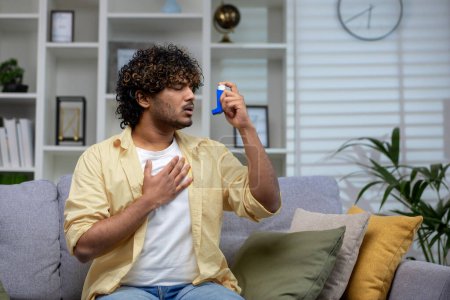 A man in a casual outfit is sitting on a sofa in a well-lit living room, holding an asthma inhaler, portraying health management at home.