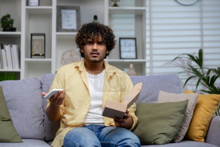 Perplexed Indian man sitting on couch with phone and open box, potentially facing online shopping scam.