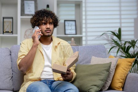 Homme indien perplexe tenant téléphone et boîte ouverte, victime potentielle de l'escroquerie du shopping en ligne, se sentant trompé à la maison.
