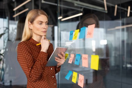 Photo for Focused businesswoman using sticky notes for brainstorming and planning on a glass wall in a modern office setting. - Royalty Free Image
