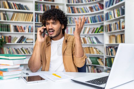 Foto de Hombre con atuendo de trabajo casual gesticulando enojado mientras habla por teléfono, sentado en un escritorio con una computadora portátil y libros. - Imagen libre de derechos