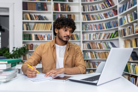 Photo for Serious young man with curly hair focused on studying using a laptop in a library setting with books around him. - Royalty Free Image
