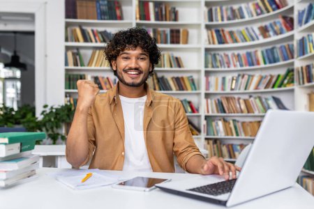 Photo for Joyful young man triumphantly clenches fist looking at laptop in library, expressing excitement for his achievement or win. - Royalty Free Image
