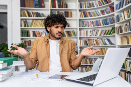 Photo for Perplexed young student sitting with a laptop in a library, surrounded by books, looking for assistance. - Royalty Free Image
