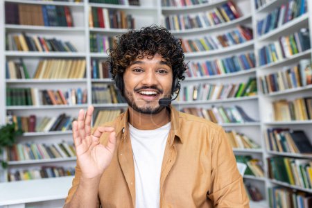 Estudiante alegre participando en el aprendizaje en línea, haciendo un gesto amistoso a la cámara mientras usa un auricular en una biblioteca.