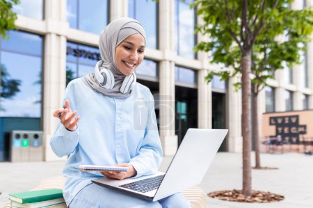 Konzentrierte junge Studentin im Hijab mit Kopfhörern, die auf ihrem Laptop lernt und sich auf dem Campus Notizen macht.