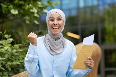 Imagen vibrante de una alegre mujer musulmana usando un hiyab, celebrando exuberantemente mientras sostiene un sobre al aire libre con un fondo verde borroso.