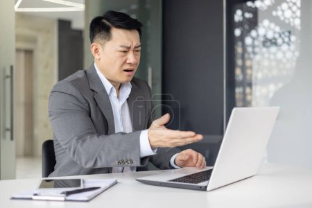 Foto de Hombre de negocios asiático en traje gris que parece confundido mientras trabaja en su computadora portátil en un espacio de trabajo de oficina moderno. - Imagen libre de derechos