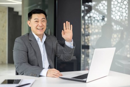 Cheerful Asian businessman in a grey suit waves while working at his laptop in a bright, contemporary office setting.