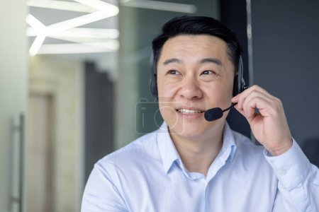 Professional Asian businessman using a headset, engaged in a conversation in a well-lit contemporary office setting.