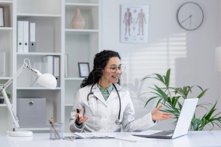 Latino femme médecin engageant une conversation par appel vidéo avec un patient dans un cadre de bureau de clinique moderne.