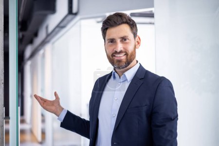 Portrait of a smiling, confident businessman in a modern office setting extending his hand as if presenting or greeting.
