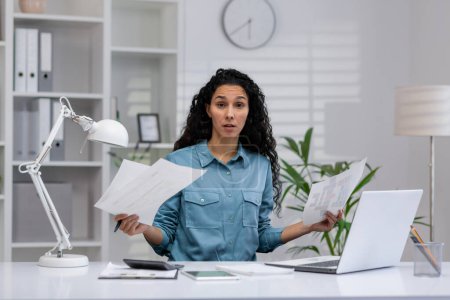 A Latina businesswoman looks confused while multitasking at her home office, surrounded by paperwork and a laptop.