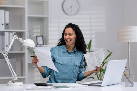Téléchargez les photos : Femme hispanique joyeuse examinant les documents avec un sourire dans son cadre bien organisé de bureau à la maison, incarnant le confort professionnel et la productivité. - en image libre de droit