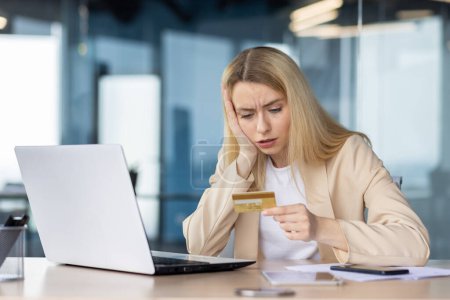 Anxious businesswoman holding a credit card and looking at laptop with concern, suspecting a fraudulent transaction at her office desk.