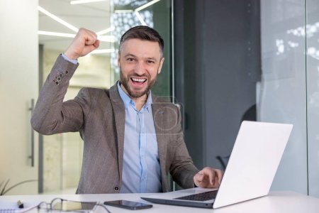 Téléchargez les photos : Un homme d'affaires triomphant lève le poing dans un geste de succès tout en travaillant sur son ordinateur portable dans un cadre de bureau moderne. - en image libre de droit