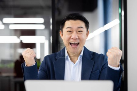 An elated businessman in a blue suit punches the air with joy in front of his laptop in a well-lit modern office.