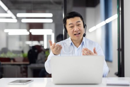 Cheerful Asian businessman conversing on a video call with a laptop and headset in an office setting.