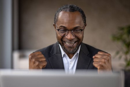 Foto de Un hombre de negocios afroamericano muy alegre celebra un acuerdo o logro exitoso mientras trabaja en su computadora portátil. - Imagen libre de derechos