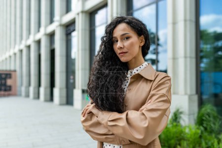 A young professional woman stands confidently outside a modern office building, exuding strength and ambition in a corporate setting.