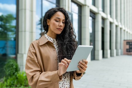 Confident professional woman browsing on a digital tablet while standing outside a contemporary office building.
