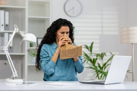 Une femme d'affaires en détresse dans une chemise bleue respire dans un sac en papier sur son lieu de travail, montrant des signes d'une attaque de panique avec un ordinateur portable devant elle.