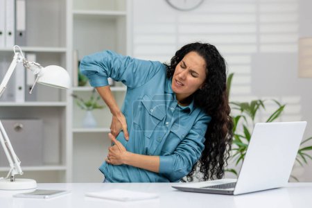 Téléchargez les photos : Une femme vêtue d'une chemise bleue ressent de fortes douleurs au dos, qui clignotent d'inconfort lorsqu'elle est assise à son bureau avec un ordinateur portable.. - en image libre de droit