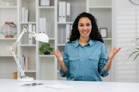 Professionelle Frau, die während eines Videogesprächs in einem gut beleuchteten Büro lächelt und gestikuliert, während sie in die Kamera blickt.
