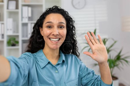 A cheerful woman is waving and smiling at the camera, depicting a friendly greeting or saying goodbye during an online video call from home.