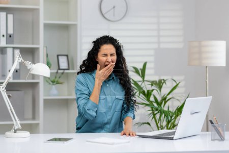Überarbeitete Geschäftsfrau mit lockigem Haar fühlt sich erschöpft und gähnt in ihrem modernen Büroraum mit Laptop und Bürobedarf.