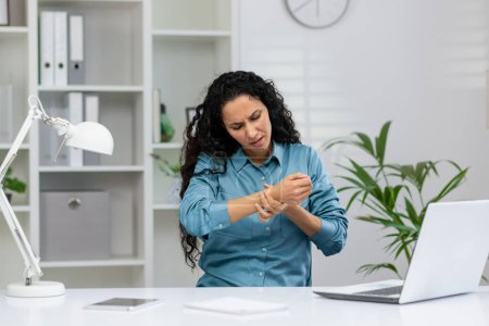 Foto de Una mujer adulta con una camisa azul siente dolor de muñeca mientras trabaja en su escritorio de la oficina, mostrando malestar y tensión. - Imagen libre de derechos