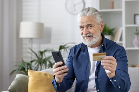 Homme âgé souriant tenant une carte de crédit et utilisant un téléphone portable dans un cadre confortable salon, illustrant les achats en ligne ou bancaires.