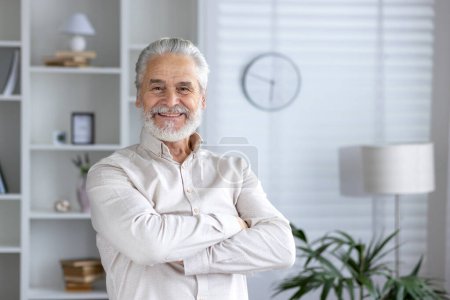 Portrait d'un homme âgé joyeux debout avec les bras croisés à la maison, rayonnant de confiance et de positivité dans un cadre confortable et élégant.