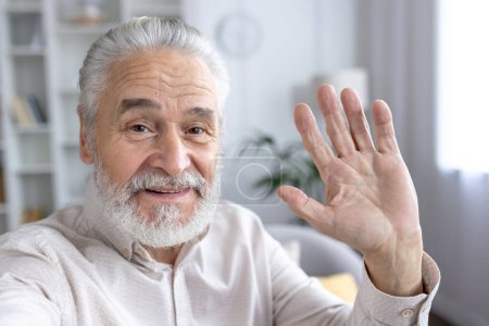 Retrato de un anciano alegre con barba, saludando a la cámara desde un espacio acogedor y luminoso.