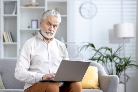 Caballero anciano enfocado navegando por Internet en su computadora portátil mientras está sentado en un sofá en una sala de estar bien iluminada.