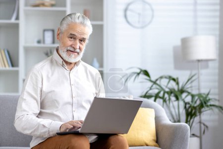 Fröhlicher älterer Herr mit Laptop, während er gemütlich in seinem gut beleuchteten Wohnzimmer zu Hause sitzt und aktives Altern präsentiert.