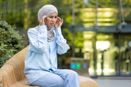 A woman wearing a hijab outdoors appears stressed and holds her head, showing signs of a headache or anxiety.